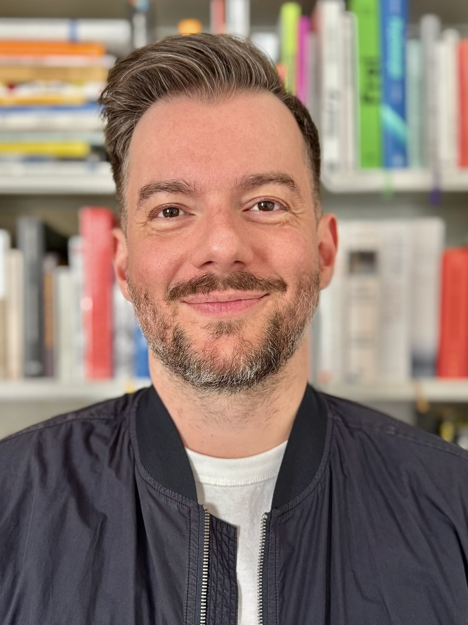 The portrait shows a man with combed back brown hair and a beard. He is wearing a dark blue jacket over a white T-shirt and is standing in front of a bookshelf filled with colorful books.